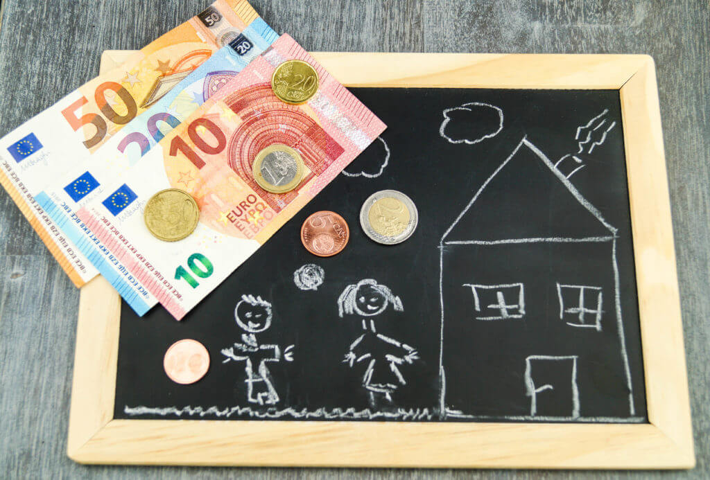 Symbolbild Baukindergeld: Euroscheine liegen neben einem Kreidebild von Kindern, die neben einem Haus stehen