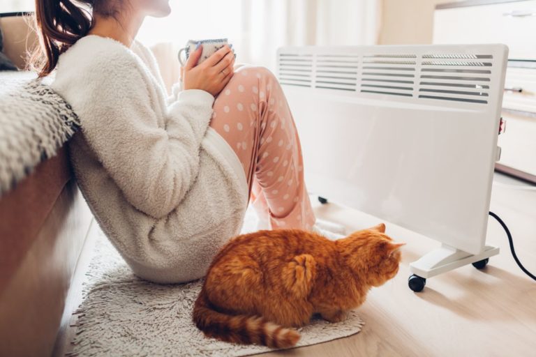 Eine Frau sitzt mit ihrer Katze vor einer Elektroheizung und hält eine Tasse