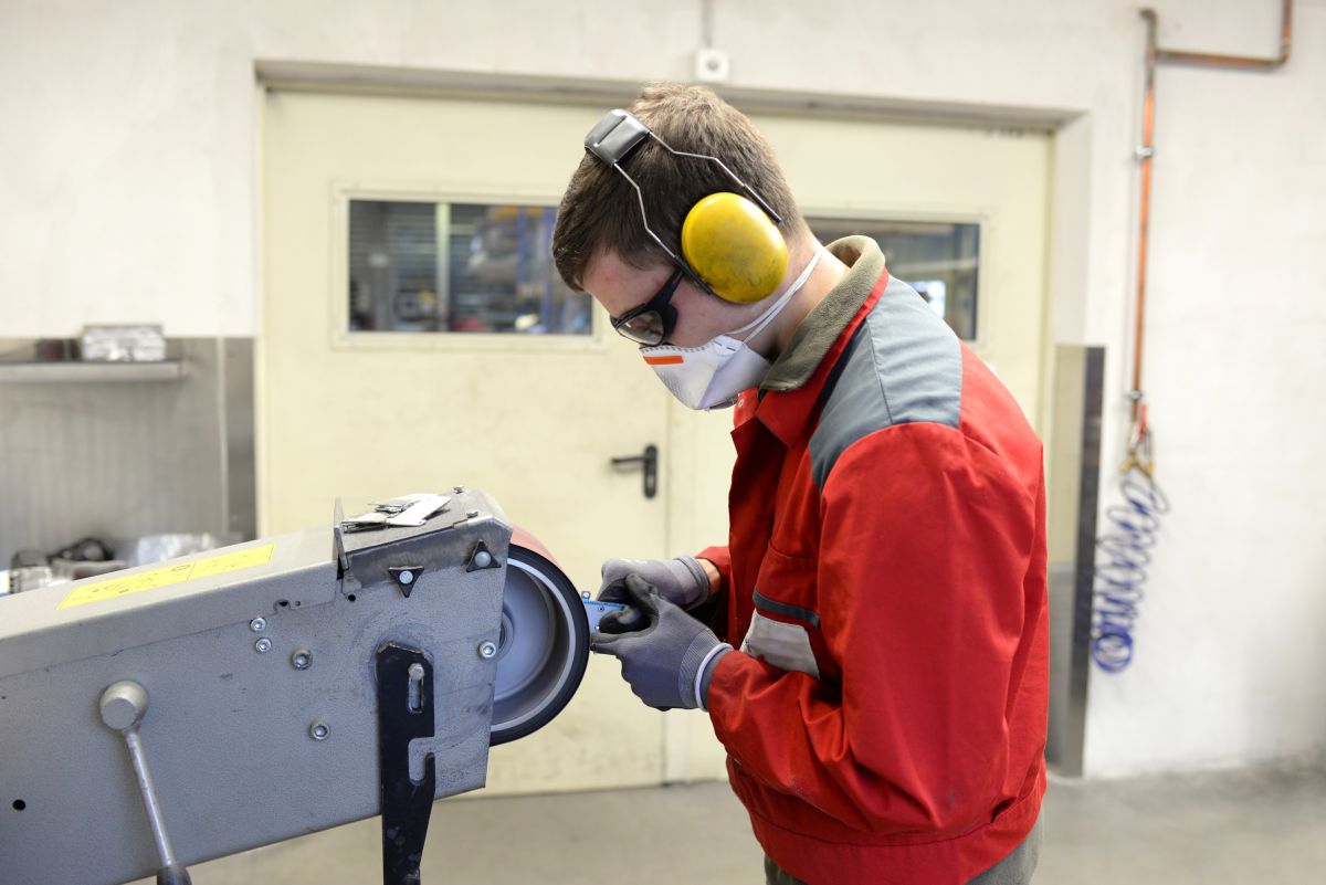 Arbeiter schleift Werkstück im Metallbau. Er trägt eine Atemschutzmaske, Gehörschutz und Handschuhe.