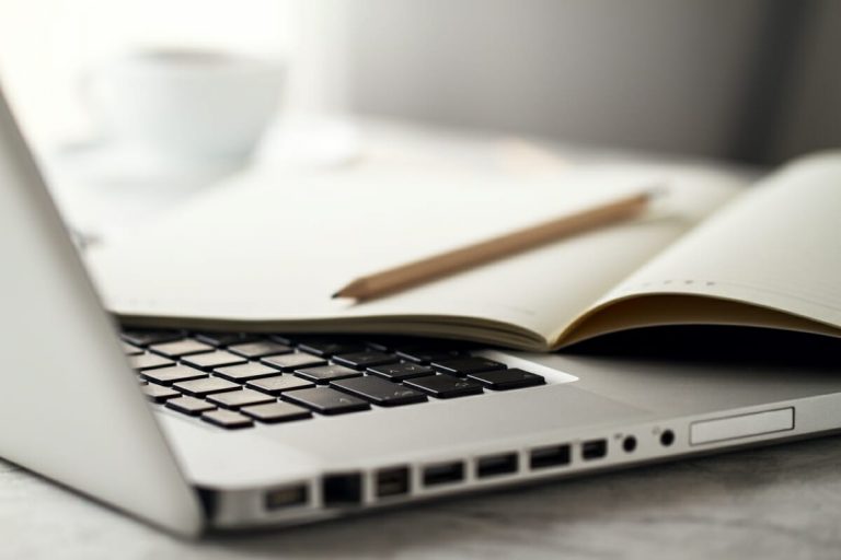 Bleifstift und Notizbuch liegen auf einem offenen Laptop. Im Hintergrund steht eine Tasse Kaffee.