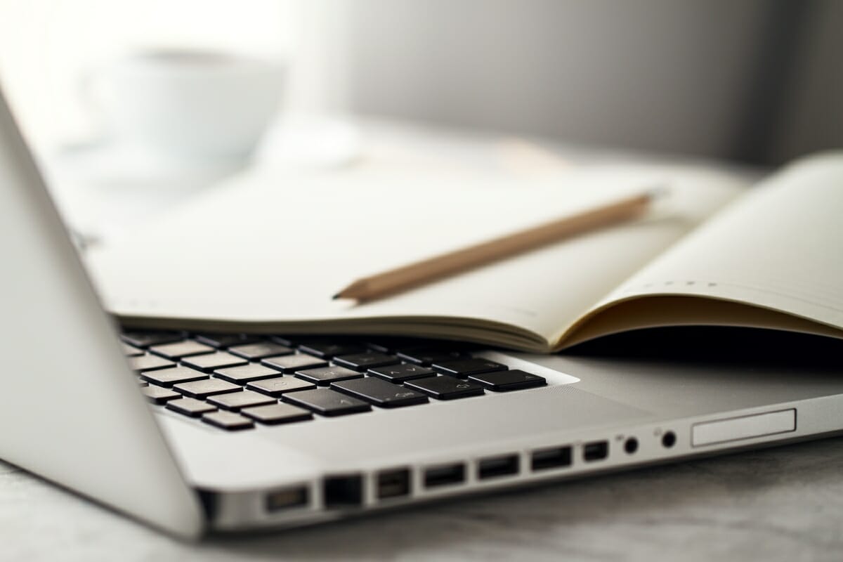 Bleifstift und Notizbuch liegen auf einem offenen Laptop. Im Hintergrund steht eine Tasse Kaffee.
