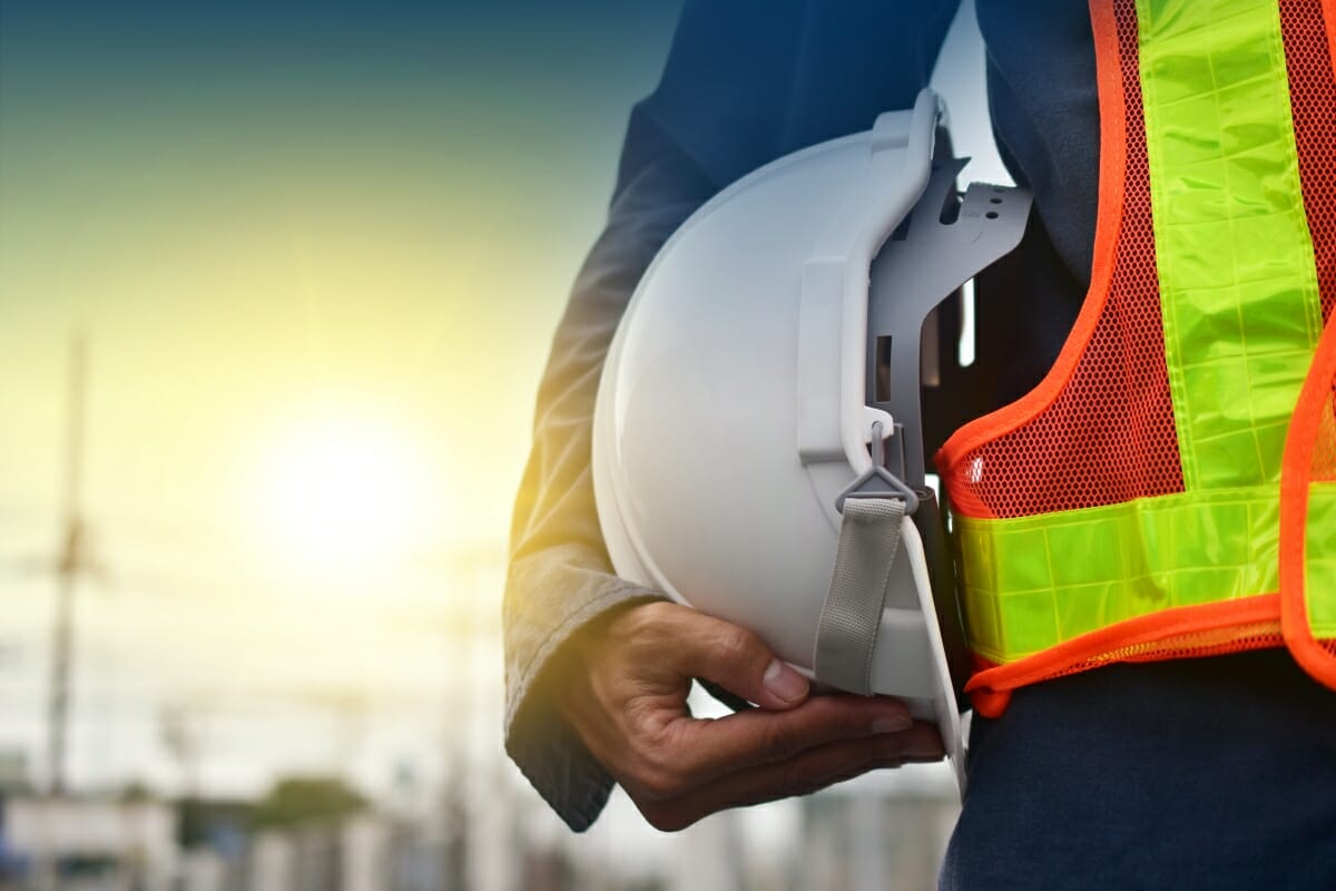 Bauarbeiter hält einen Schutzhelm und trägt eine Warnweste auf einer Baustelle. Im Hintergrund scheint die Sonne.