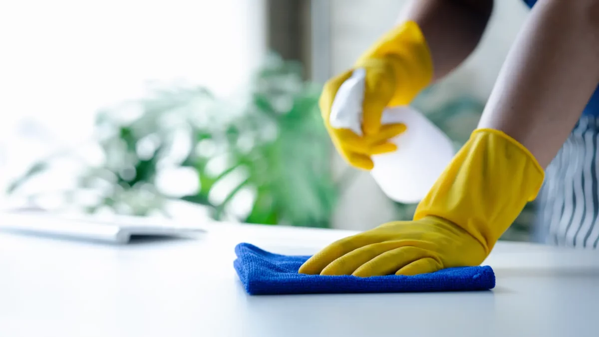 Person reinigt einen Büroschreibtisch mit einem Putztuch. Sie trägt gelbe Handschuhe und sprüht Reinigungsmittel auf den Tisch.