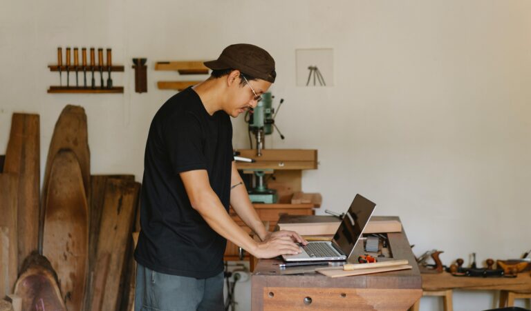 A craftsman focuses on his laptop in a woodworking workshop, blending technology and traditional craft.