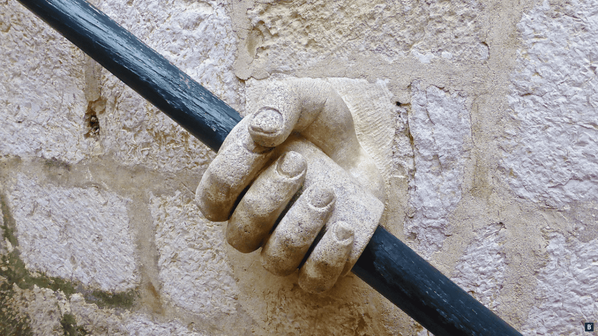 Ein alter Holzhandlauf, der durch eine Steinhand gehalten wird.